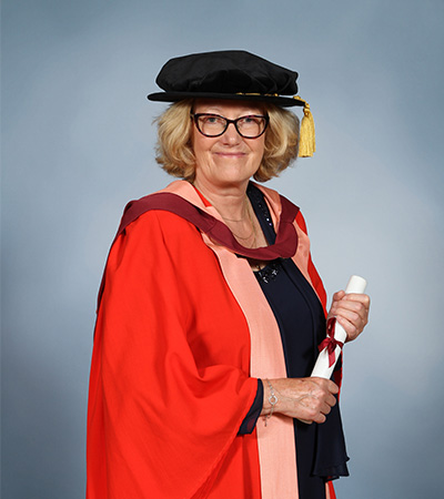An image of Joanna Holmes wearing a red Honorary Degree gown smiling and holding her degree scroll
